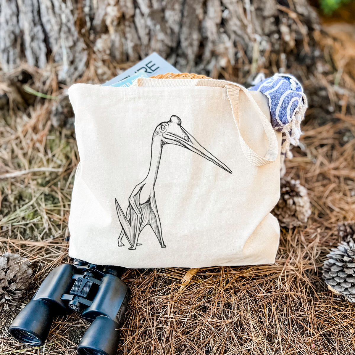 Canvas tote bag printed with a black ink illustration of a Quetzalcoatlus pterosaur standing on all fours, resting on dry grass beside a pair of binoculars.