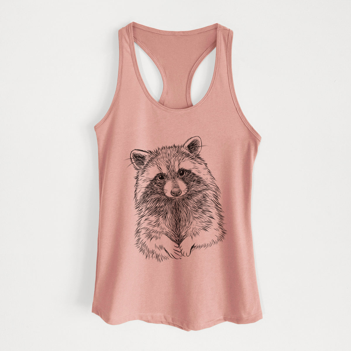 Desert pink women's racerback tank top laid flat, printed with a detailed black ink portrait of a raccoon with its front paws resting in front of it.