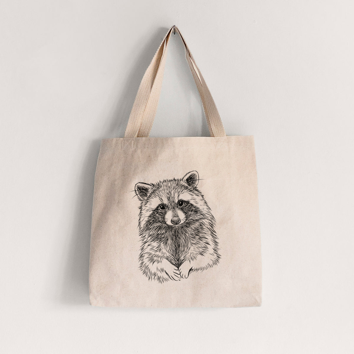 Natural canvas tote bag printed with a detailed black ink portrait of a raccoon with its front paws resting in front of it.