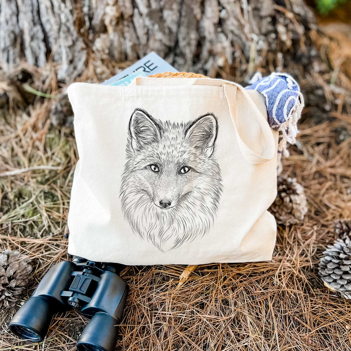 Canvas tote bag printed with a detailed black ink sketch of a red fox's face looking straight ahead, its fur drawn in fine pencil-like strokes, resting on dry grass beside a pair of binoculars.