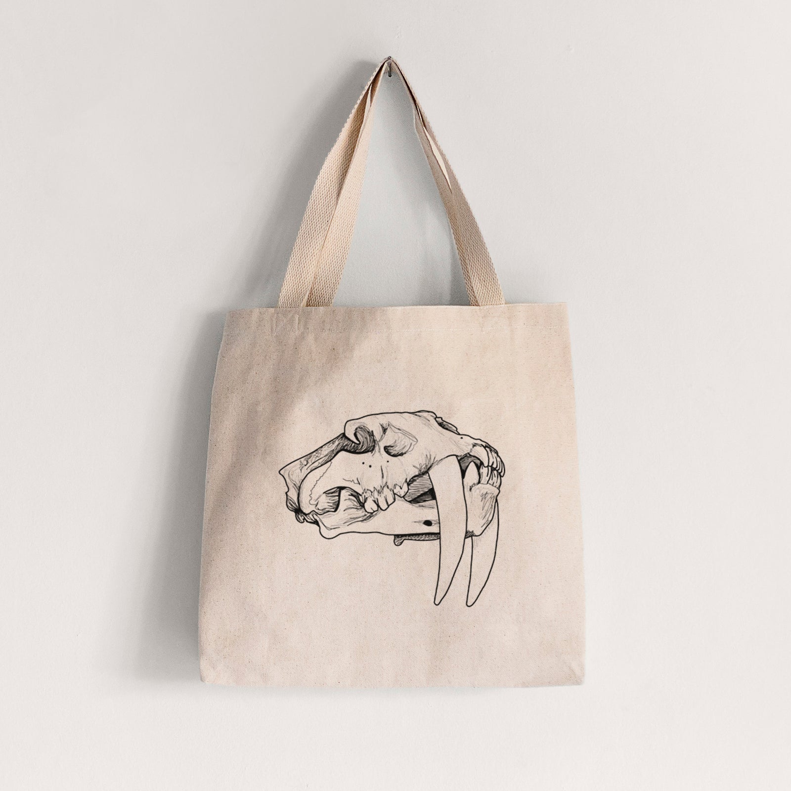 Natural canvas tote bag printed with a detailed black ink illustration of a saber-toothed tiger skull.