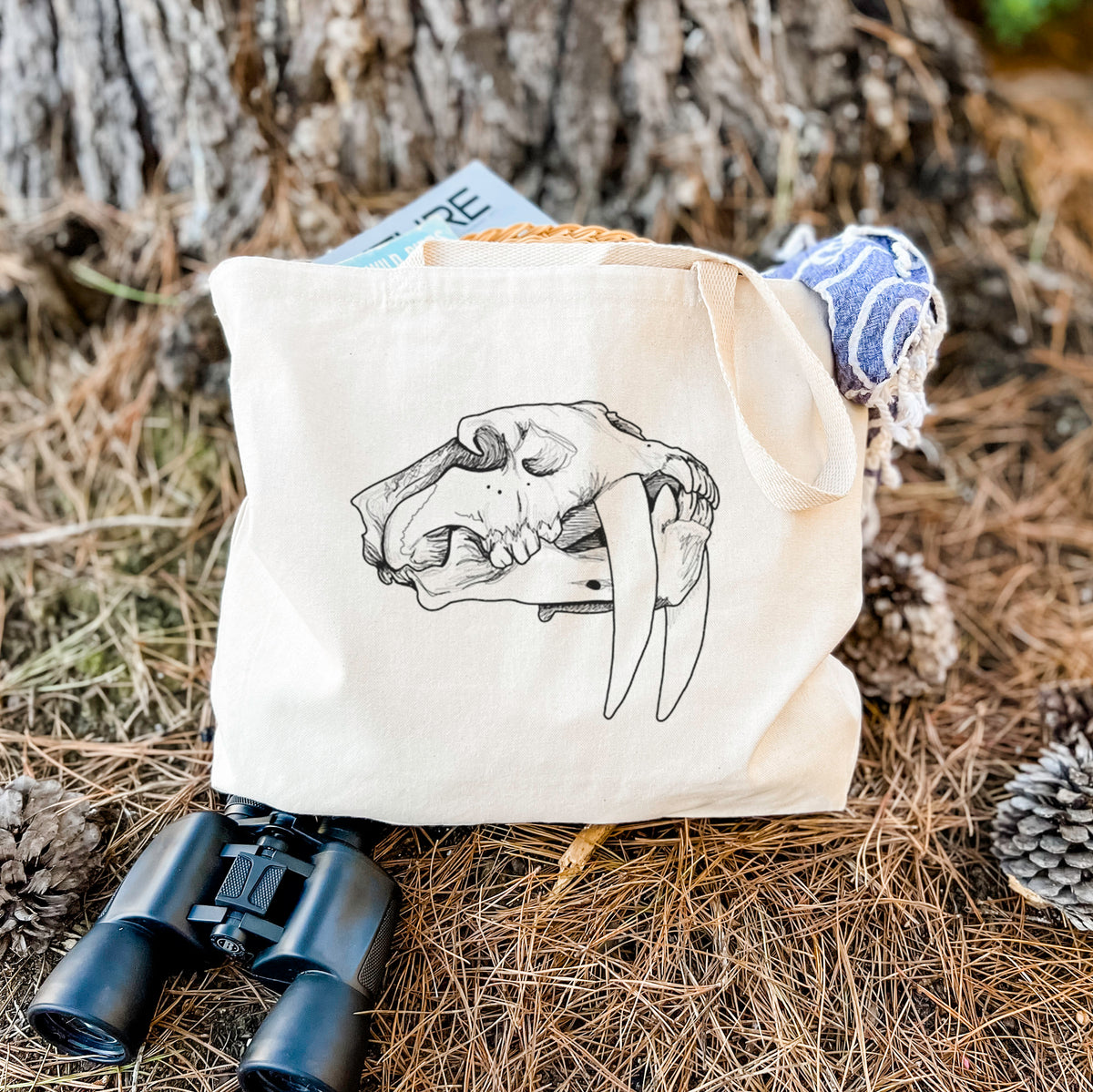 Canvas tote bag printed with a detailed black ink illustration of a saber-toothed tiger skull, resting on dry grass beside a pair of binoculars.