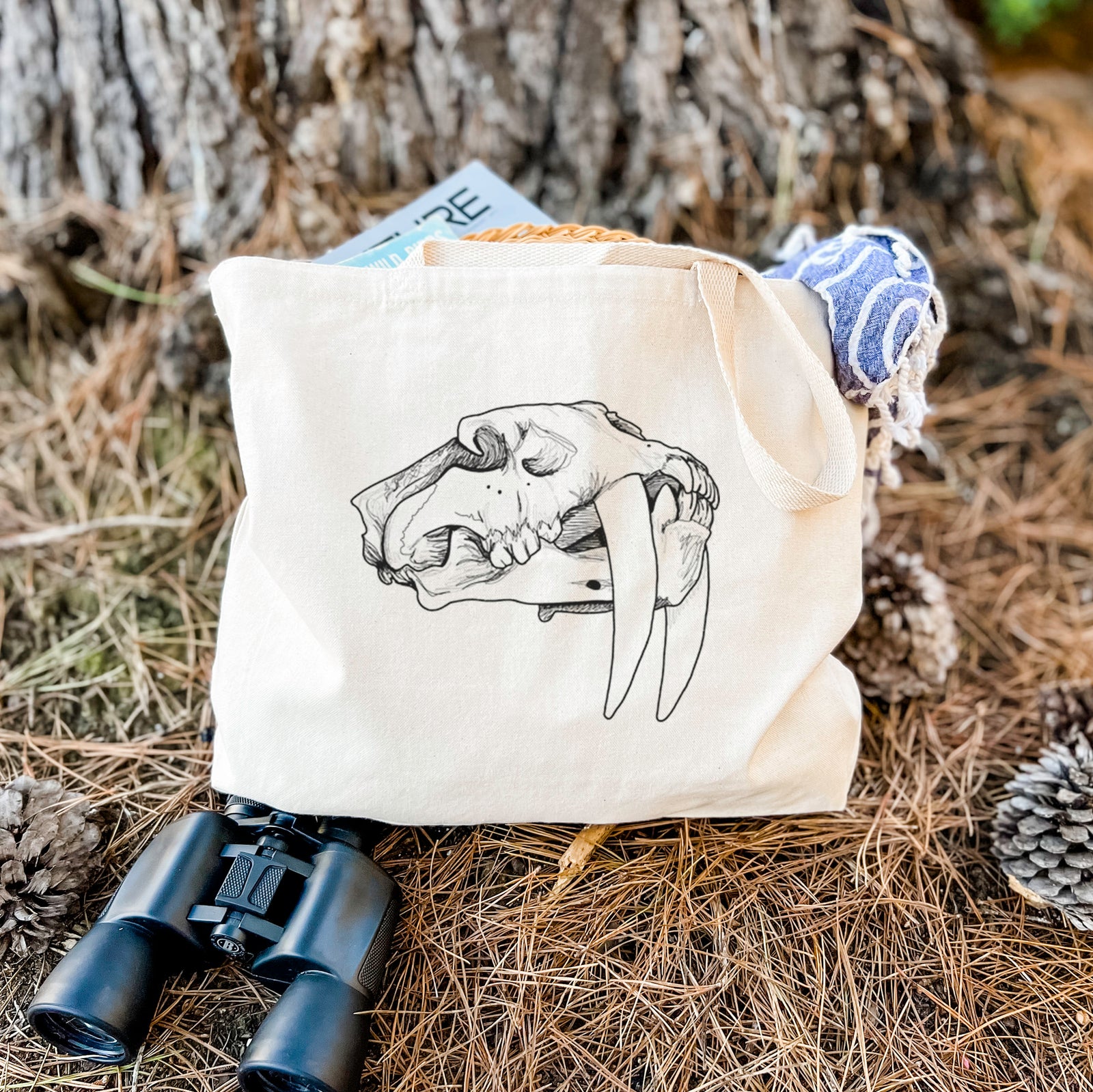 Natural canvas tote bag printed with a detailed black ink illustration of a saber-toothed tiger skull.