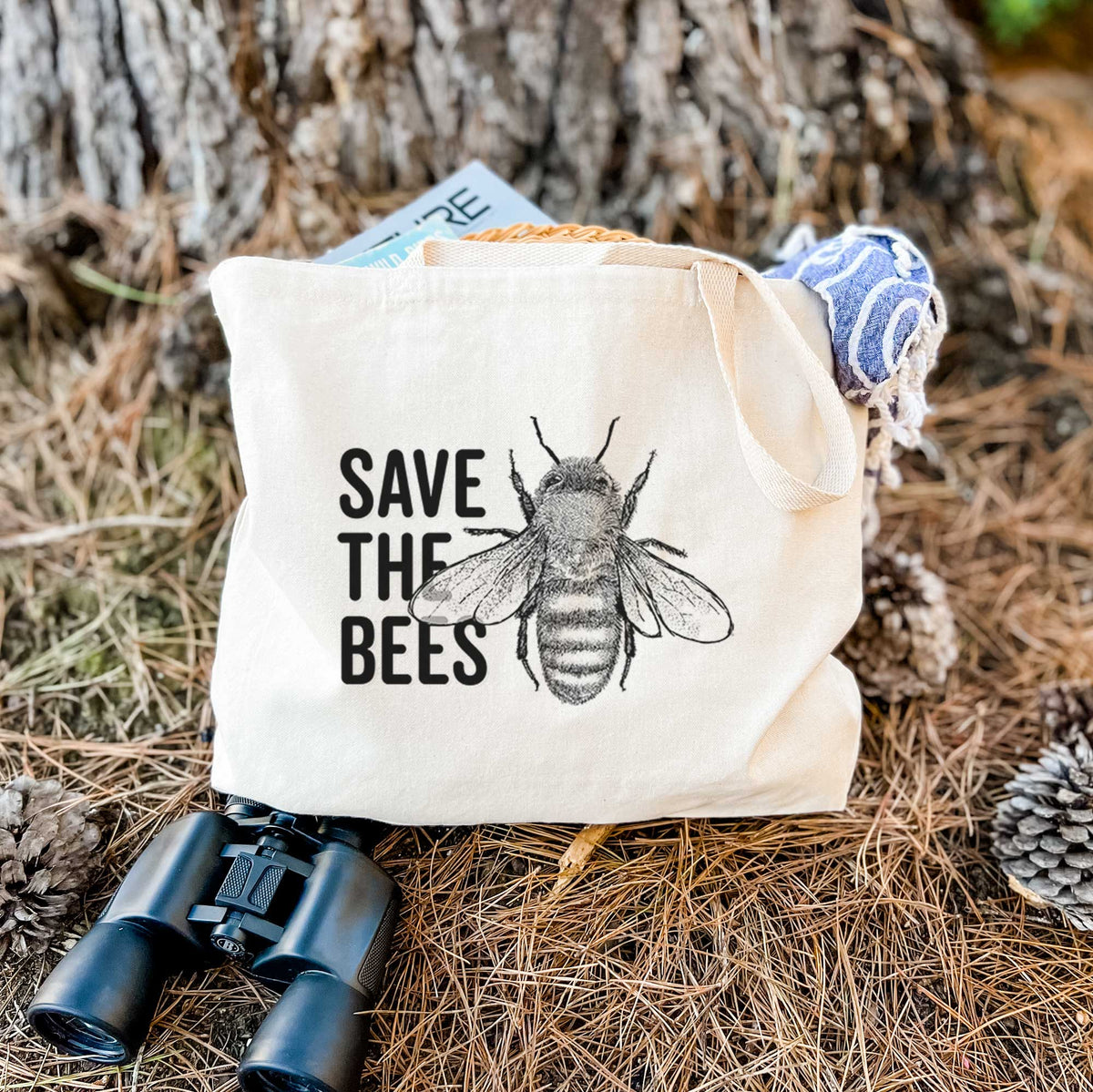 Canvas tote bag printed with bold black words reading Save the Bees beside a detailed honey bee illustration, resting on dry grass beside a pair of binoculars.