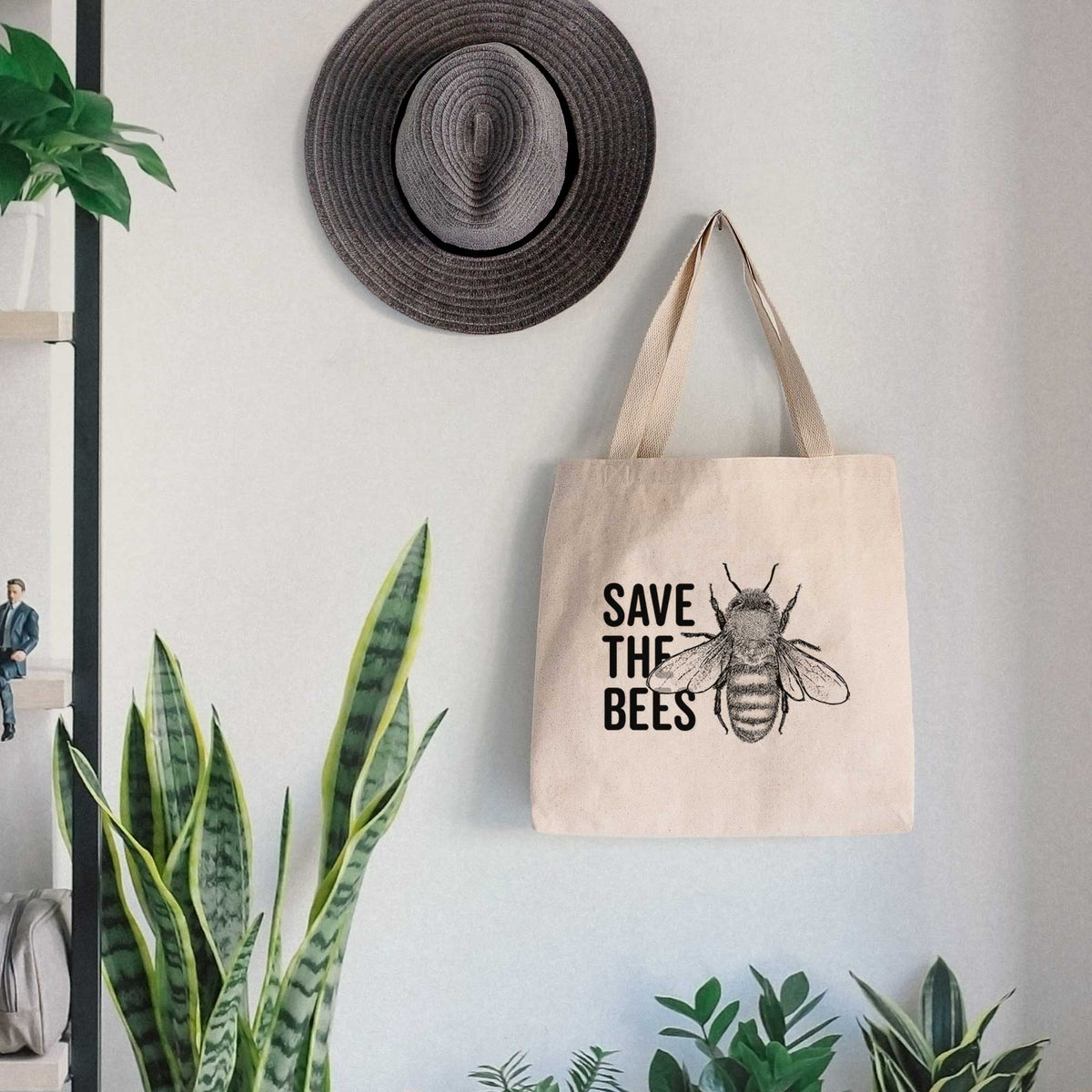 Canvas tote bag printed with bold black words reading Save the Bees beside a detailed honey bee illustration, hanging on a wall hook below a hat, next to potted plants.