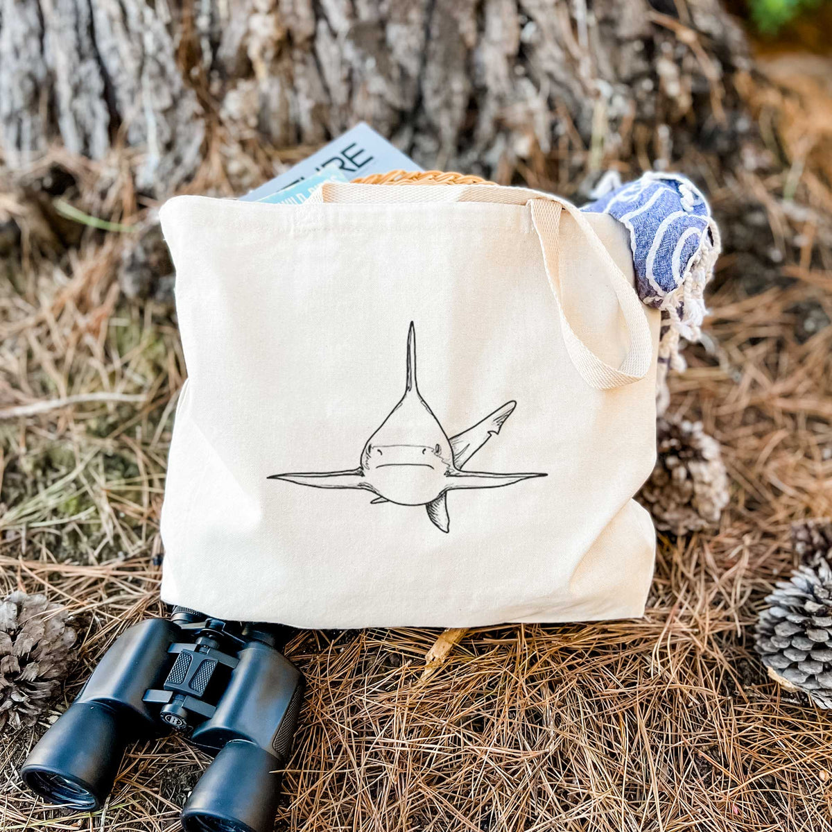Canvas tote bag printed with a black ink illustration of a silvertip shark swimming head-on, resting on dry grass beside a pair of binoculars.