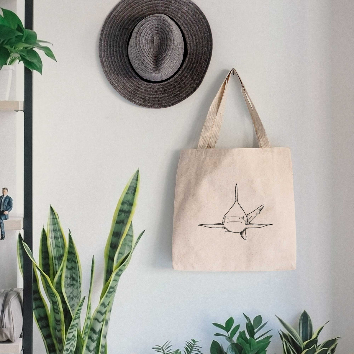 Canvas tote bag printed with a black ink illustration of a silvertip shark swimming head-on, hanging on a wall hook below a hat, next to potted plants.