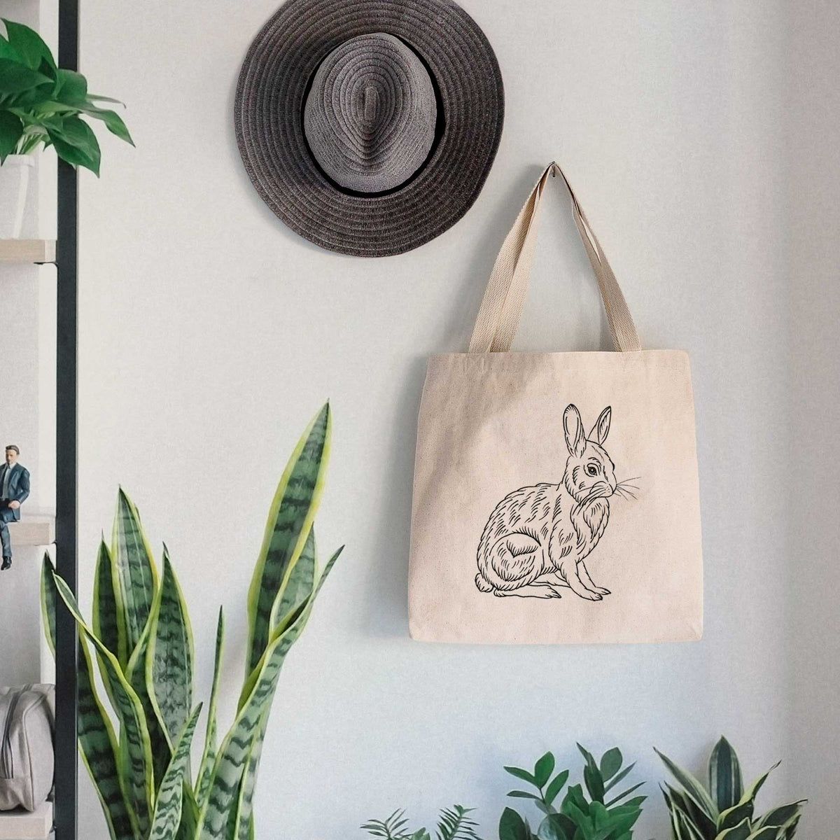 Canvas tote bag printed with a detailed black ink illustration of a snowshoe hare sitting upright, hanging on a wall hook below a hat, next to potted plants.