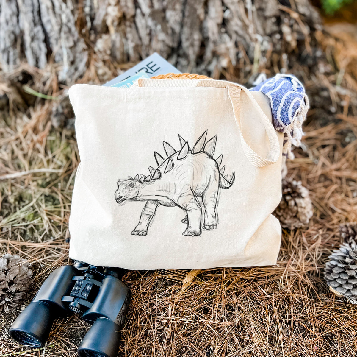 Canvas tote bag printed with a black ink illustration of a Stegosaurus with plates along its back, resting on dry grass beside a pair of binoculars.