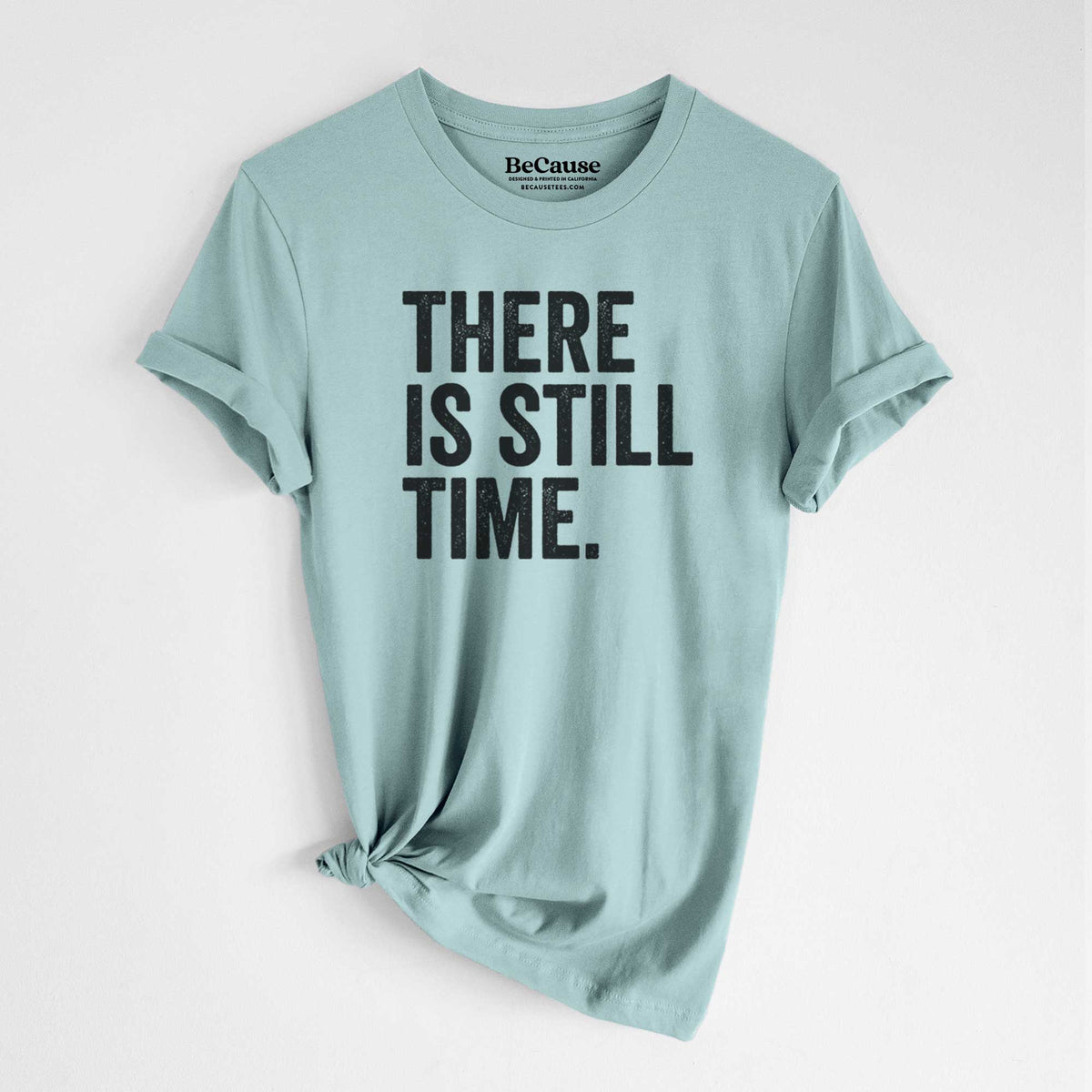 Dusty blue lightweight cotton t-shirt laid flat with the sleeves rolled, printed with bold black words reading There Is Still Time.