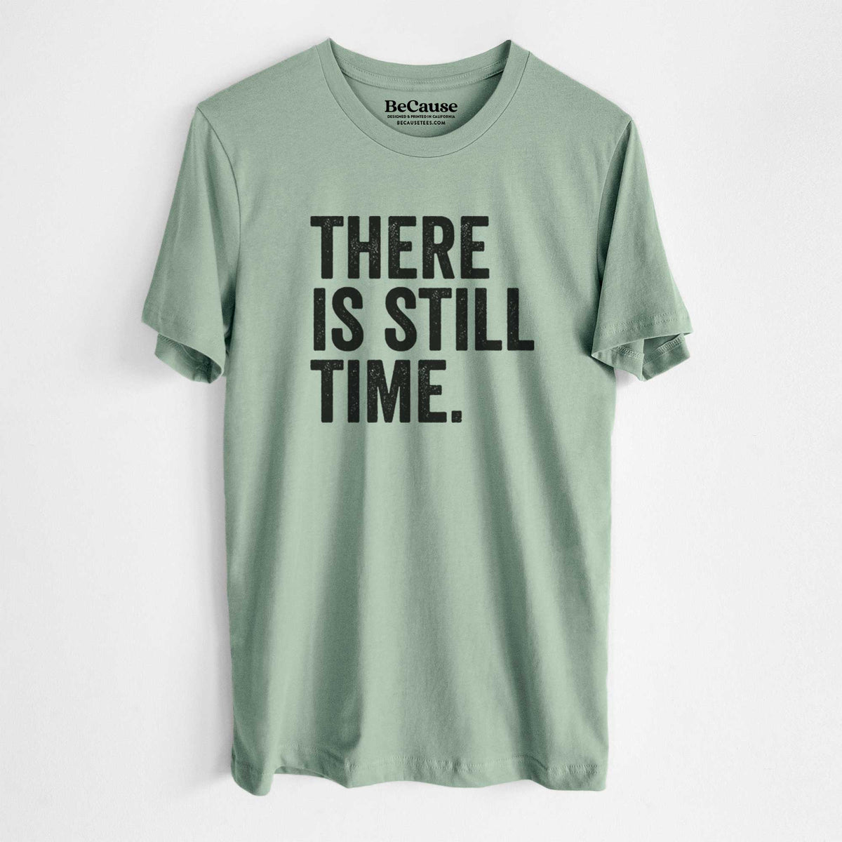 Sage green lightweight cotton t-shirt laid flat, printed with bold black words reading There Is Still Time.
