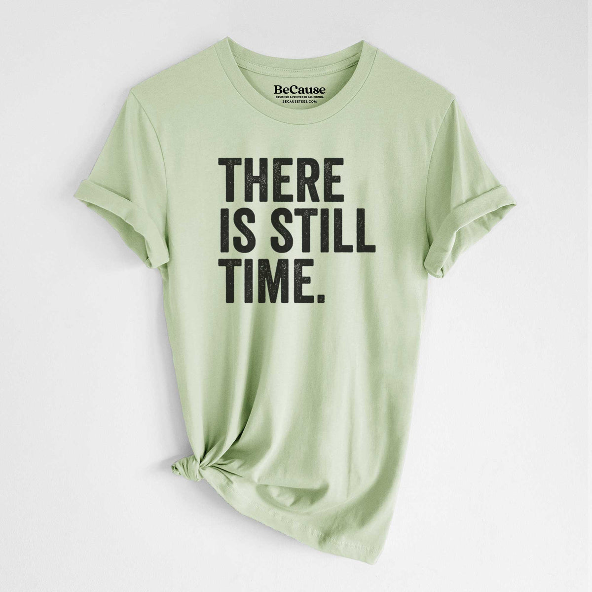 Spring green lightweight cotton t-shirt laid flat with the sleeves rolled, printed with bold black words reading There Is Still Time.