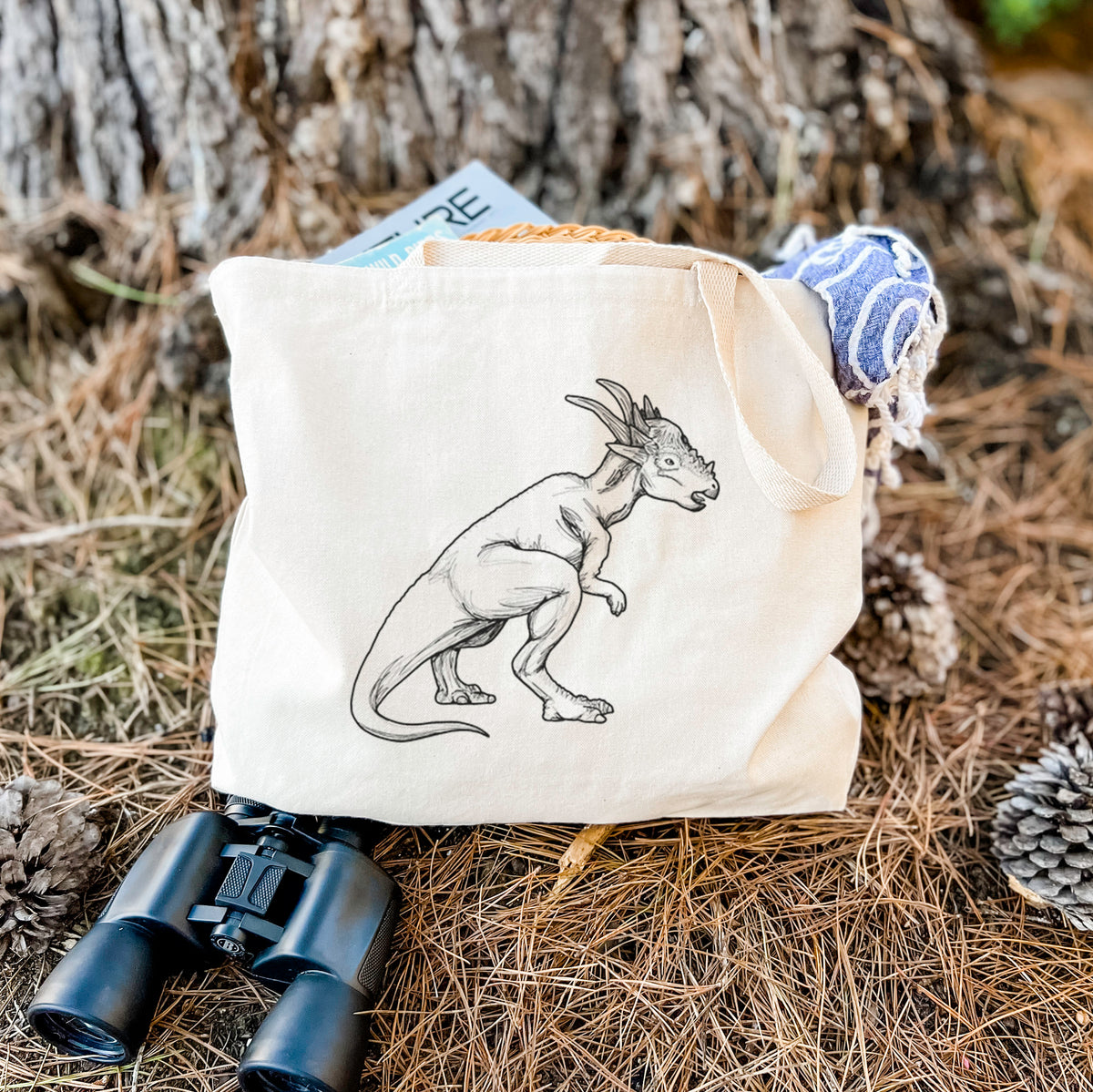 Canvas tote bag printed with a black ink illustration of a Stygimoloch dinosaur with a spiky domed head, resting on dry grass beside a pair of binoculars.