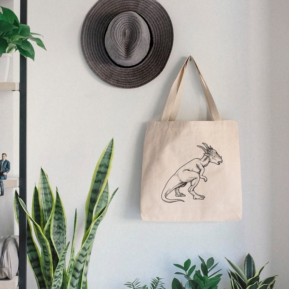 Canvas tote bag printed with a black ink illustration of a Stygimoloch dinosaur with a spiky domed head, hanging on a wall hook below a hat, next to potted plants.
