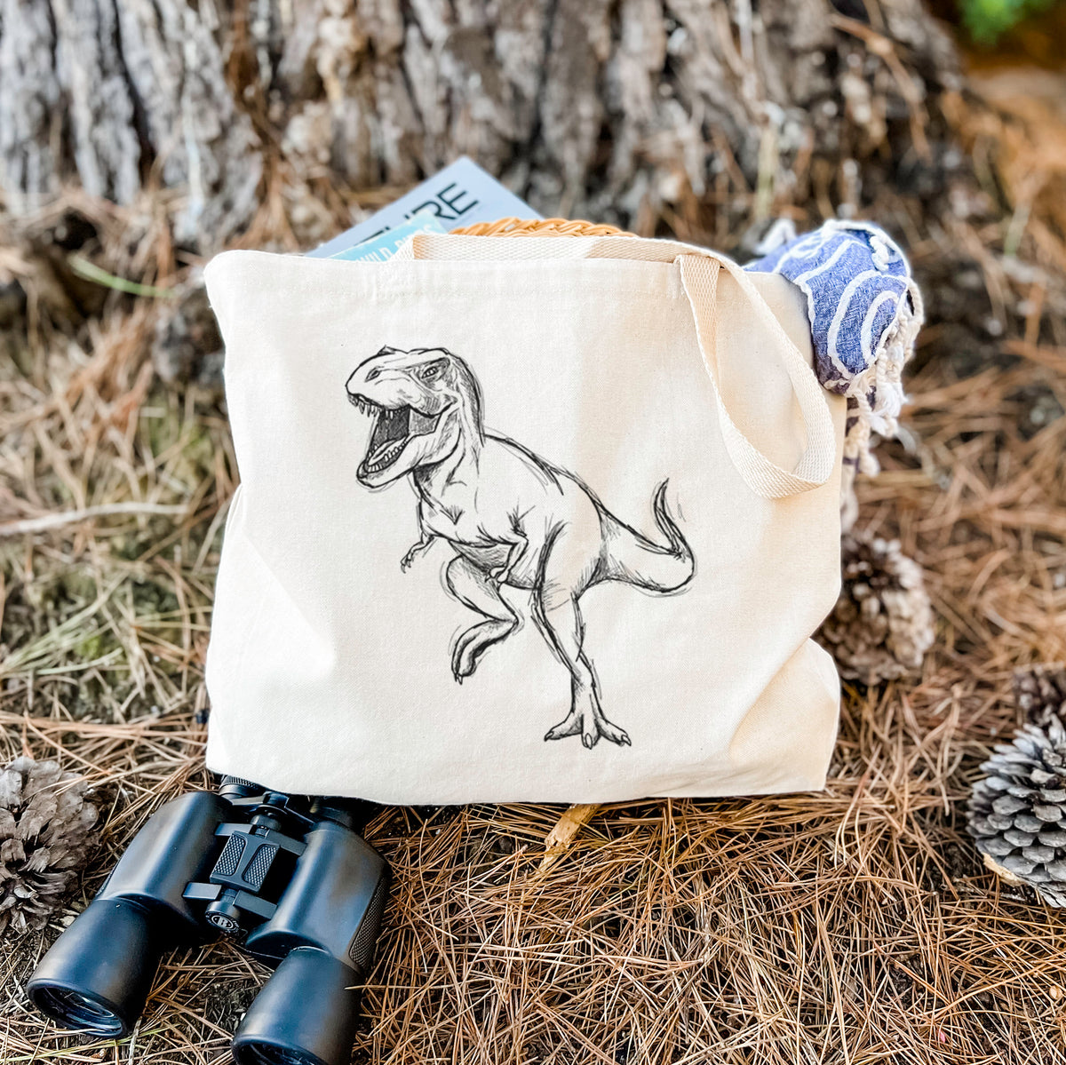 Canvas tote bag printed with a black ink illustration of a Tyrannosaurus rex roaring, resting on dry grass beside a pair of binoculars.