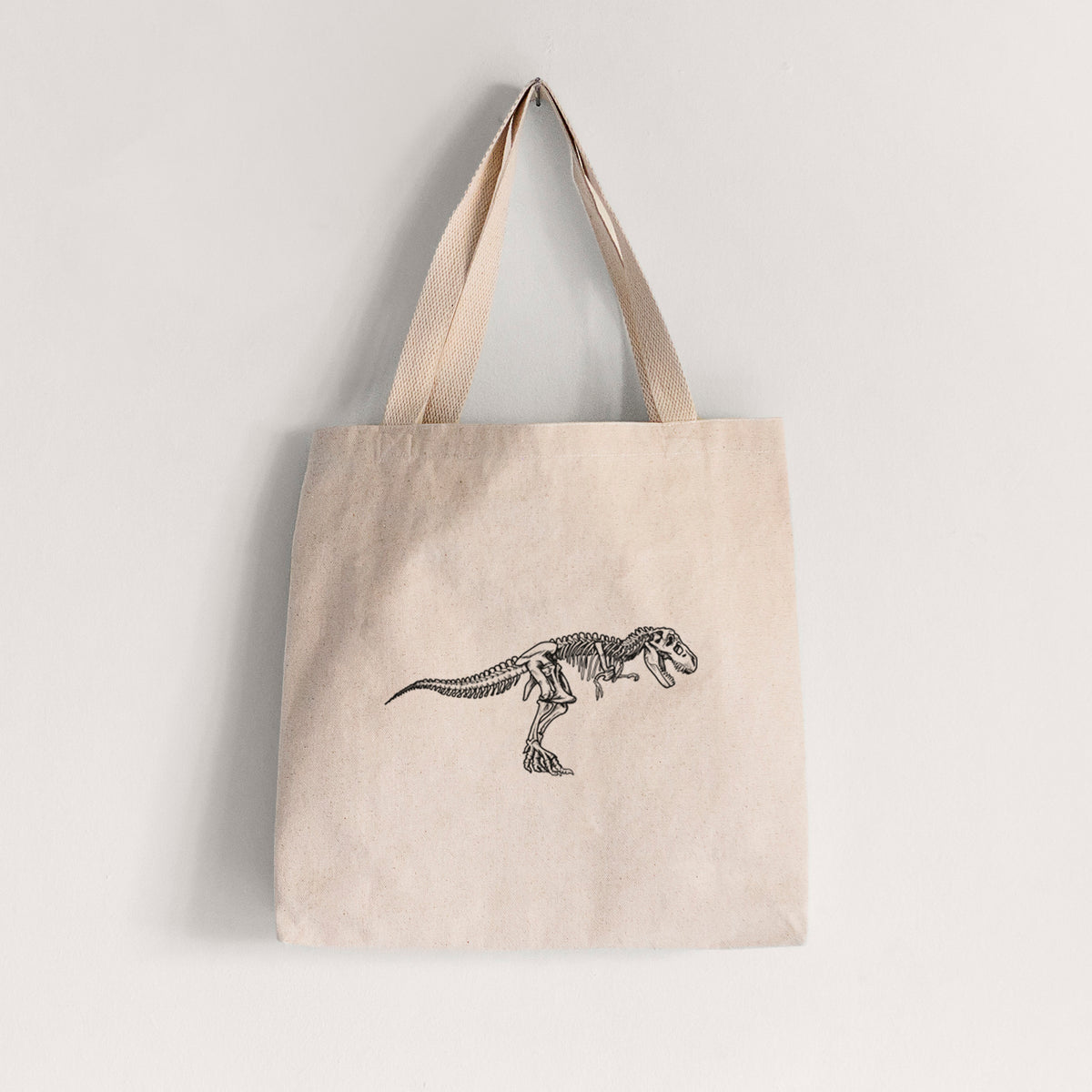Natural canvas tote bag printed with a black ink illustration of a Tyrannosaurus rex skeleton.