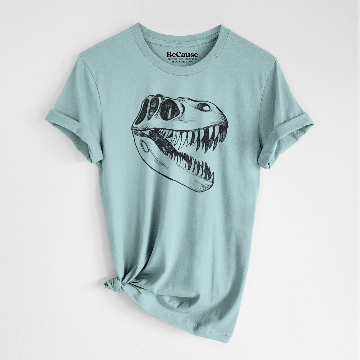 Dusty blue lightweight cotton t-shirt laid flat with the sleeves rolled, printed with a black ink illustration of a Tyrannosaurus rex skull with sharp teeth.