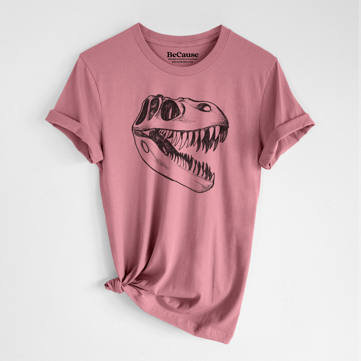 Mauve pink lightweight cotton t-shirt laid flat with the sleeves rolled, printed with a black ink illustration of a Tyrannosaurus rex skull with sharp teeth.
