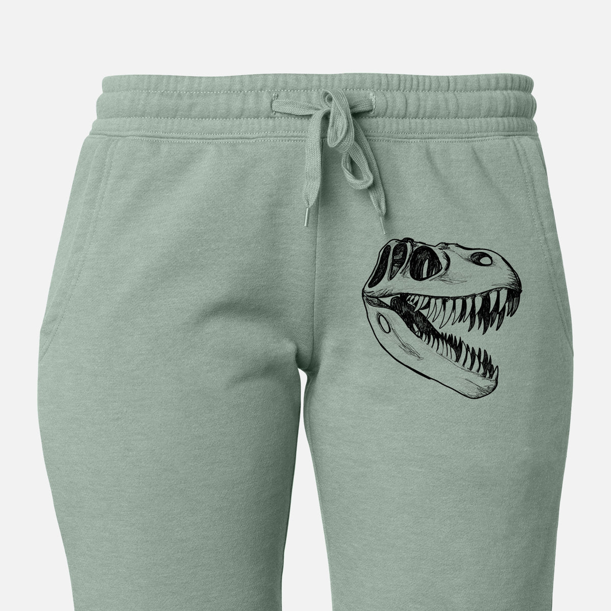 Front of sage green Cali Wave sweatpants, printed with a black ink illustration of a Tyrannosaurus rex skull with sharp teeth on the leg.