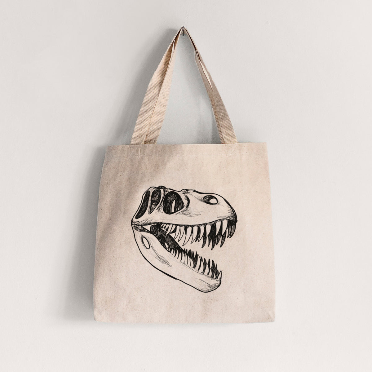 Natural canvas tote bag printed with a black ink illustration of a Tyrannosaurus rex skull with sharp teeth.