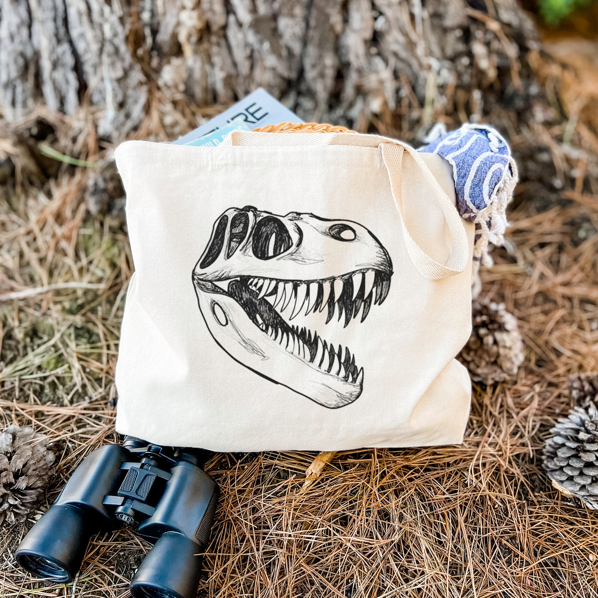 Canvas tote bag printed with a black ink illustration of a Tyrannosaurus rex skull with sharp teeth, resting on dry grass beside a pair of binoculars.