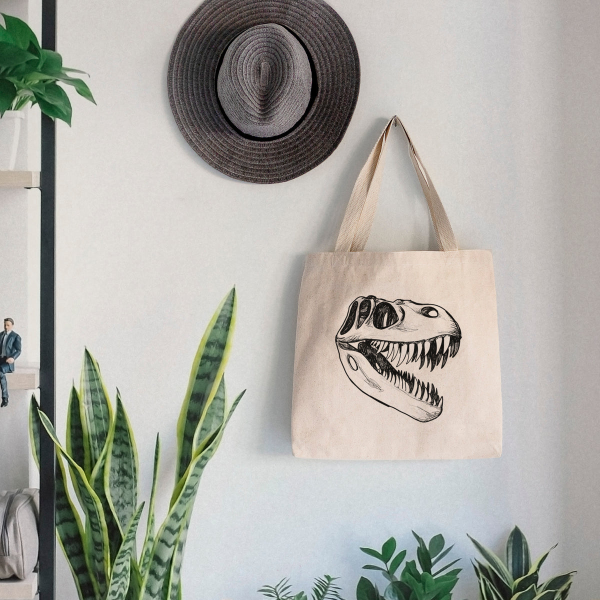 Canvas tote bag printed with a black ink illustration of a Tyrannosaurus rex skull with sharp teeth, hanging on a wall hook below a hat, next to potted plants.