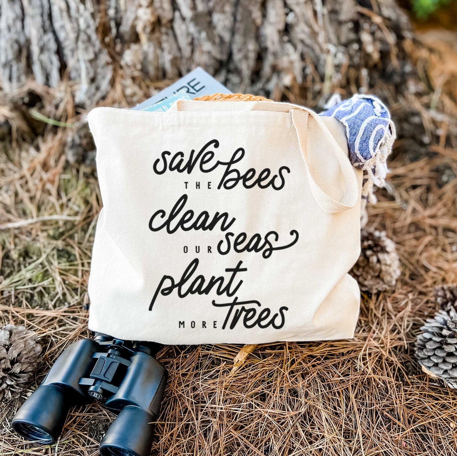 Natural canvas tote bag printed with black hand-lettered words reading Save the Bees, Clean Our Seas, Plant More Trees.