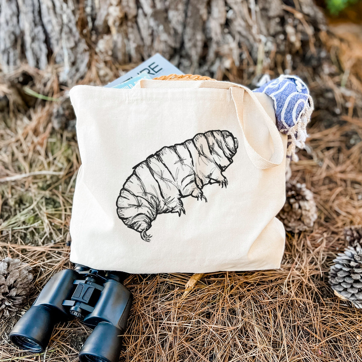 Canvas tote bag printed with a detailed black ink illustration of a tardigrade, or water bear, resting on dry grass beside a pair of binoculars.