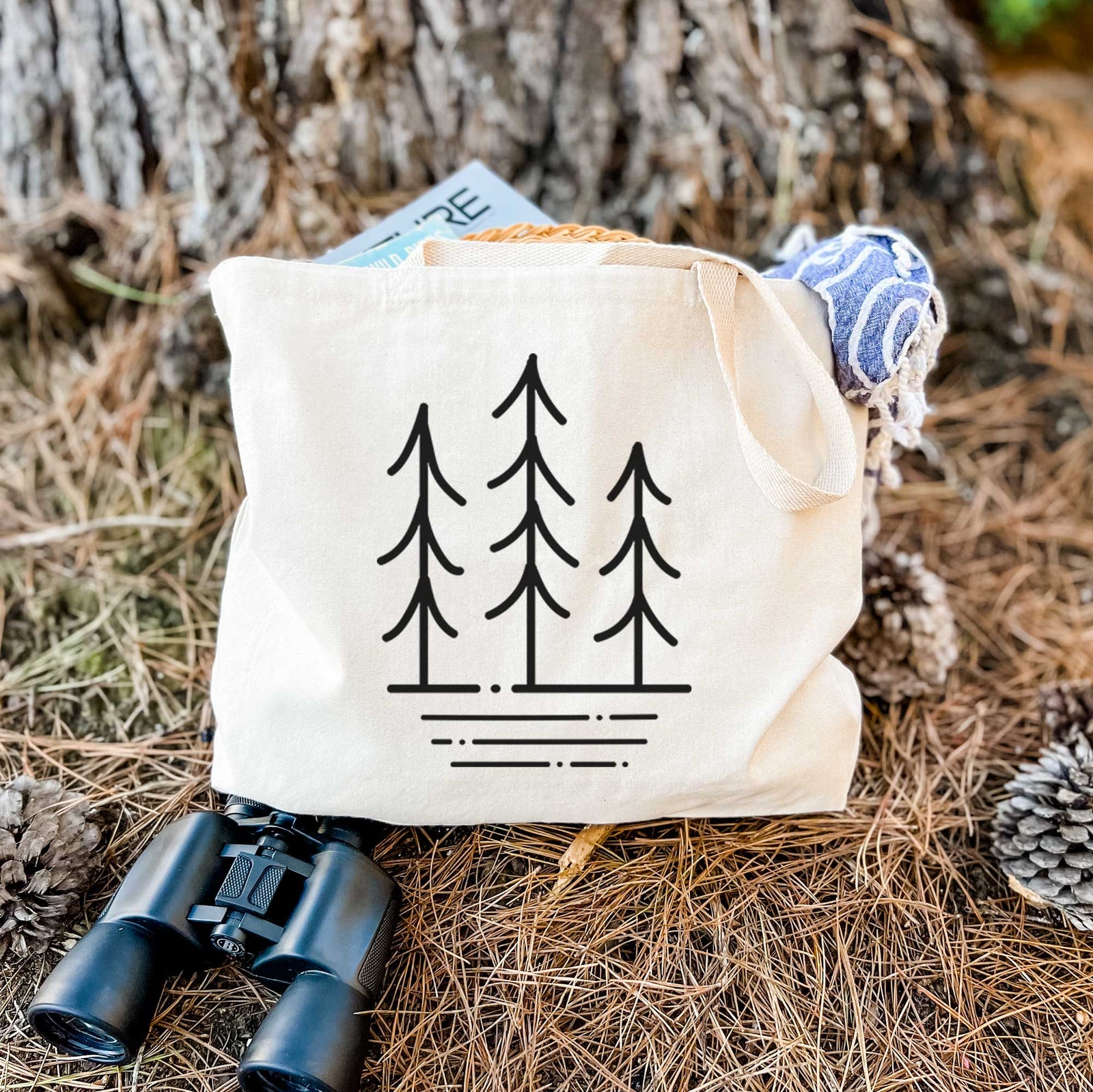 Natural canvas tote bag printed with black line art of three simple pine trees above horizontal lines.