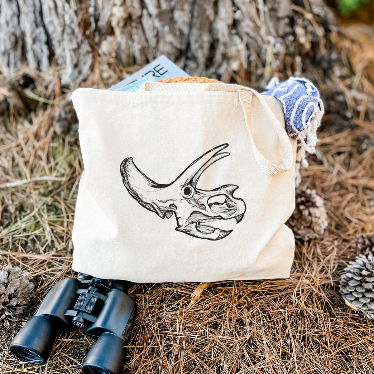 Canvas tote bag printed with a detailed black ink illustration of a Triceratops skull with its horns and frill, resting on dry grass beside a pair of binoculars.