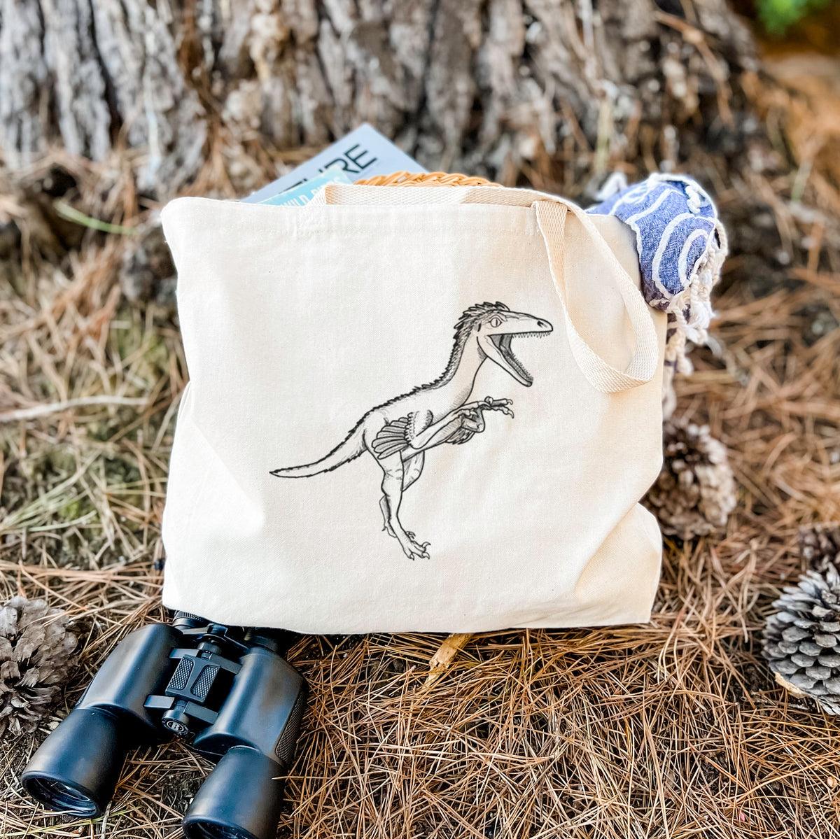Canvas tote bag printed with a black ink illustration of a Troodon dinosaur running, resting on dry grass beside a pair of binoculars.