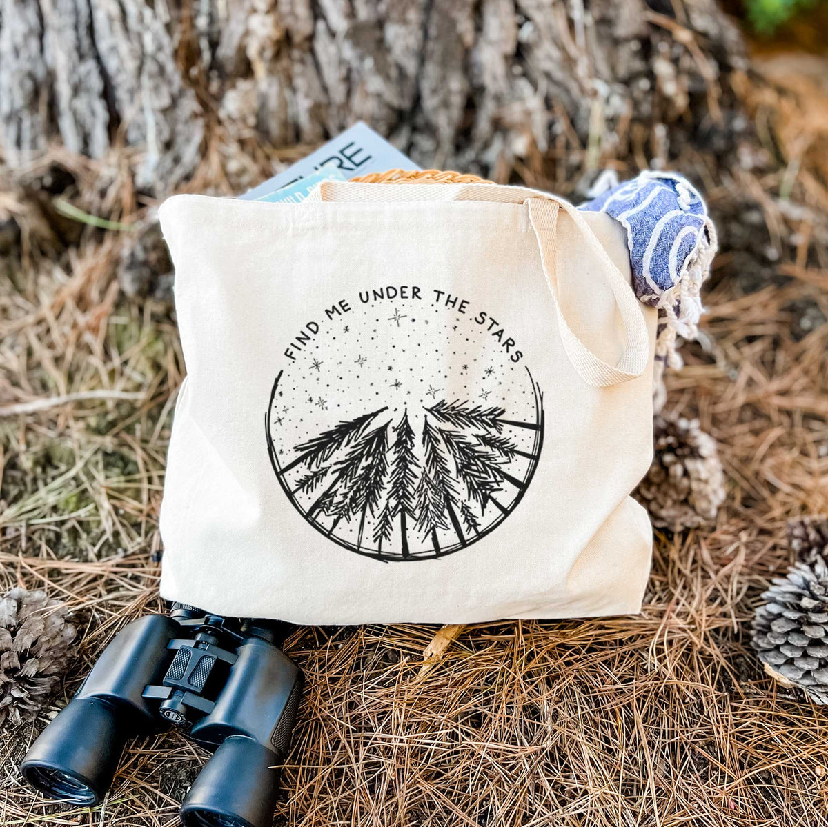 Canvas tote bag printed with a circle of pine trees under a starry sky, with the words Find Me Under the Stars, resting on dry grass beside a pair of binoculars.