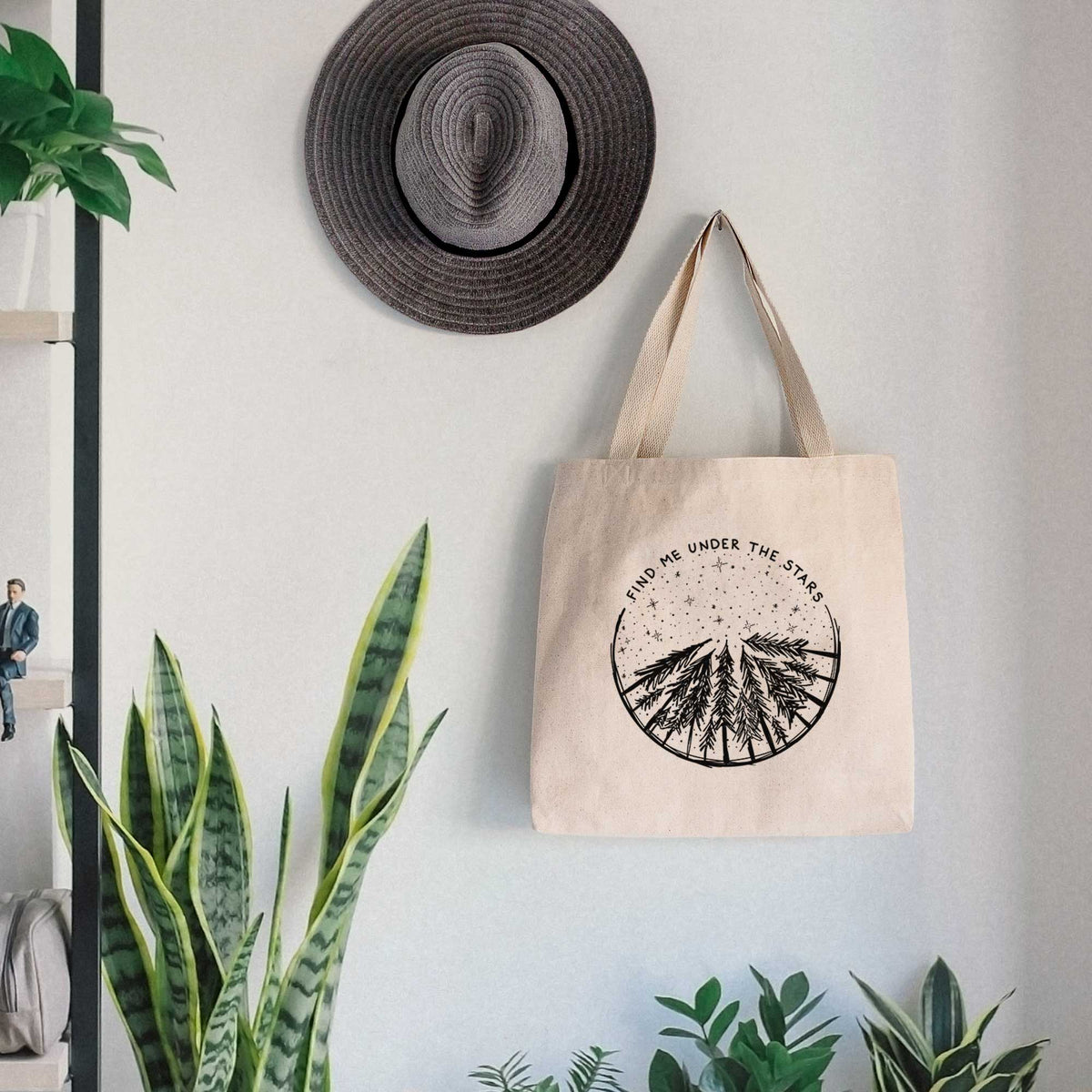 Canvas tote bag printed with a circle of pine trees under a starry sky, with the words Find Me Under the Stars, hanging on a wall hook below a hat, next to potted plants.