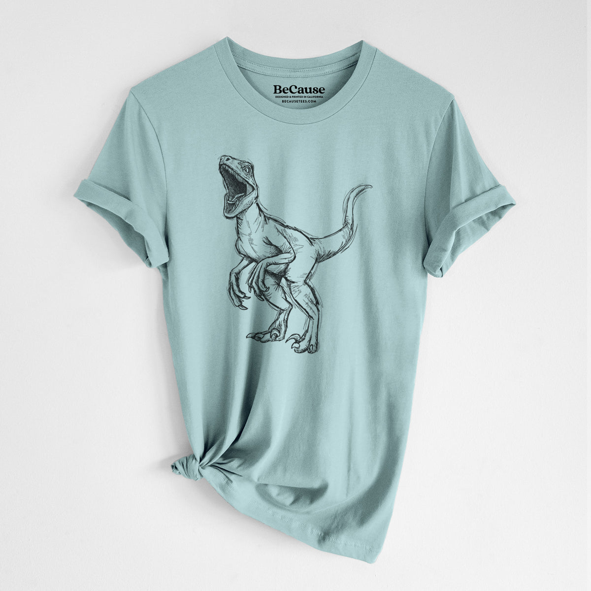 Dusty blue lightweight cotton t-shirt laid flat with the sleeves rolled, printed with a black ink illustration of a Velociraptor roaring.