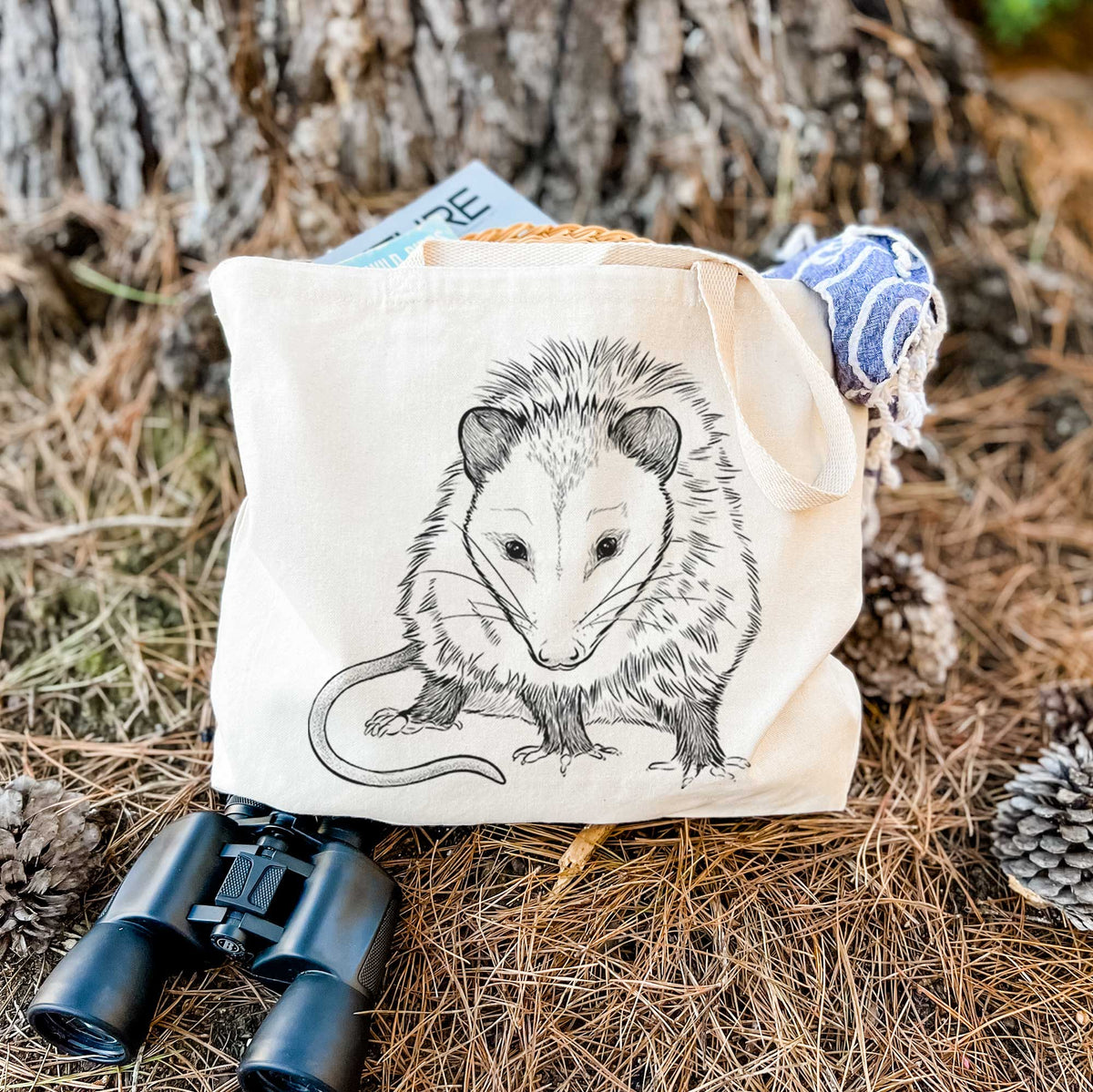 Canvas tote bag printed with a detailed black ink illustration of a Virginia opossum standing with its long tail curled, resting on dry grass beside a pair of binoculars.