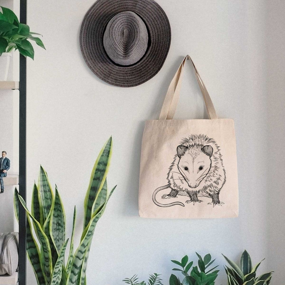 Canvas tote bag printed with a detailed black ink illustration of a Virginia opossum standing with its long tail curled, hanging on a wall hook below a hat, next to potted plants.