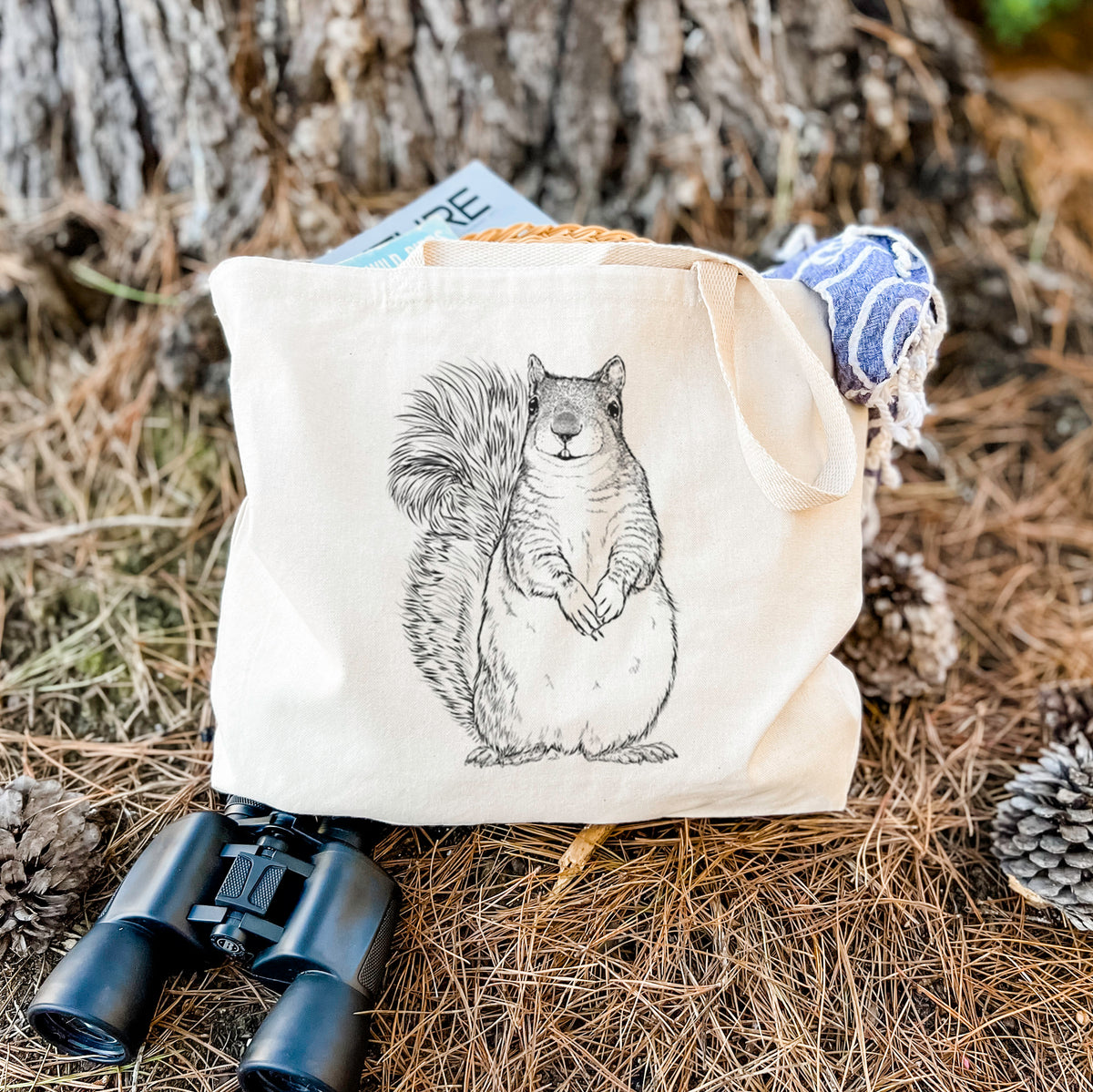Canvas tote bag printed with a detailed black ink illustration of a western grey squirrel sitting upright with a bushy tail, resting on dry grass beside a pair of binoculars.