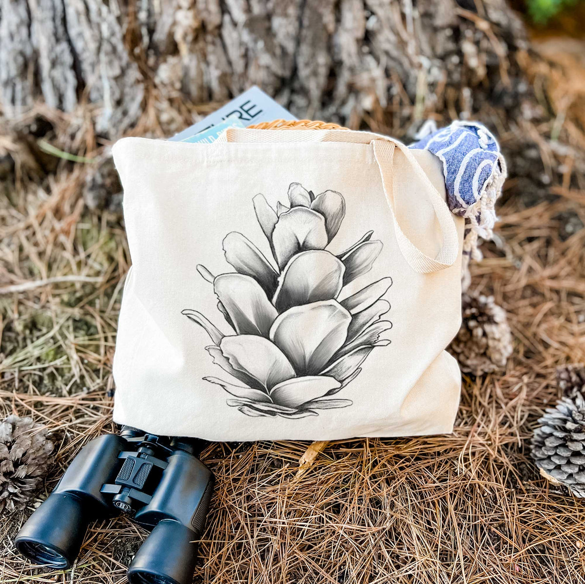 Canvas tote bag printed with a detailed black ink illustration of a western hemlock pine cone, resting on dry grass beside a pair of binoculars.