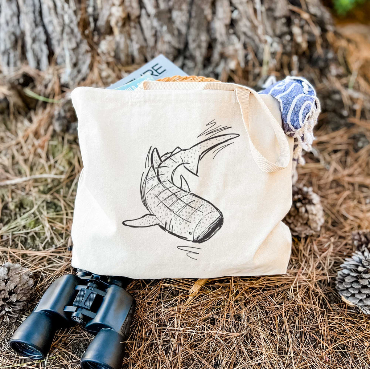Canvas tote bag printed with a black ink illustration of a spotted whale shark swimming, resting on dry grass beside a pair of binoculars.