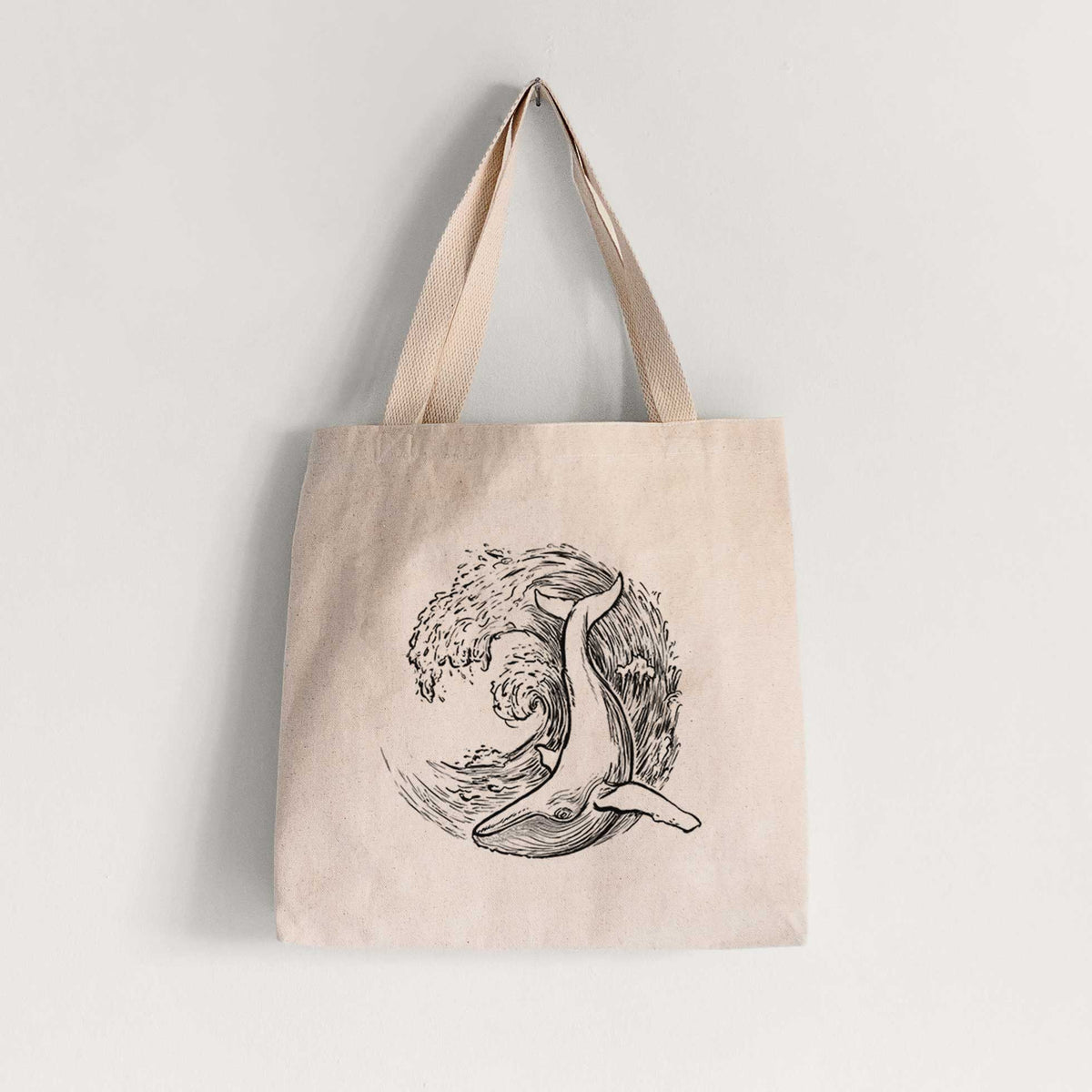 Natural canvas tote bag printed with a black ink illustration of a humpback whale swimming inside a curling ocean wave.