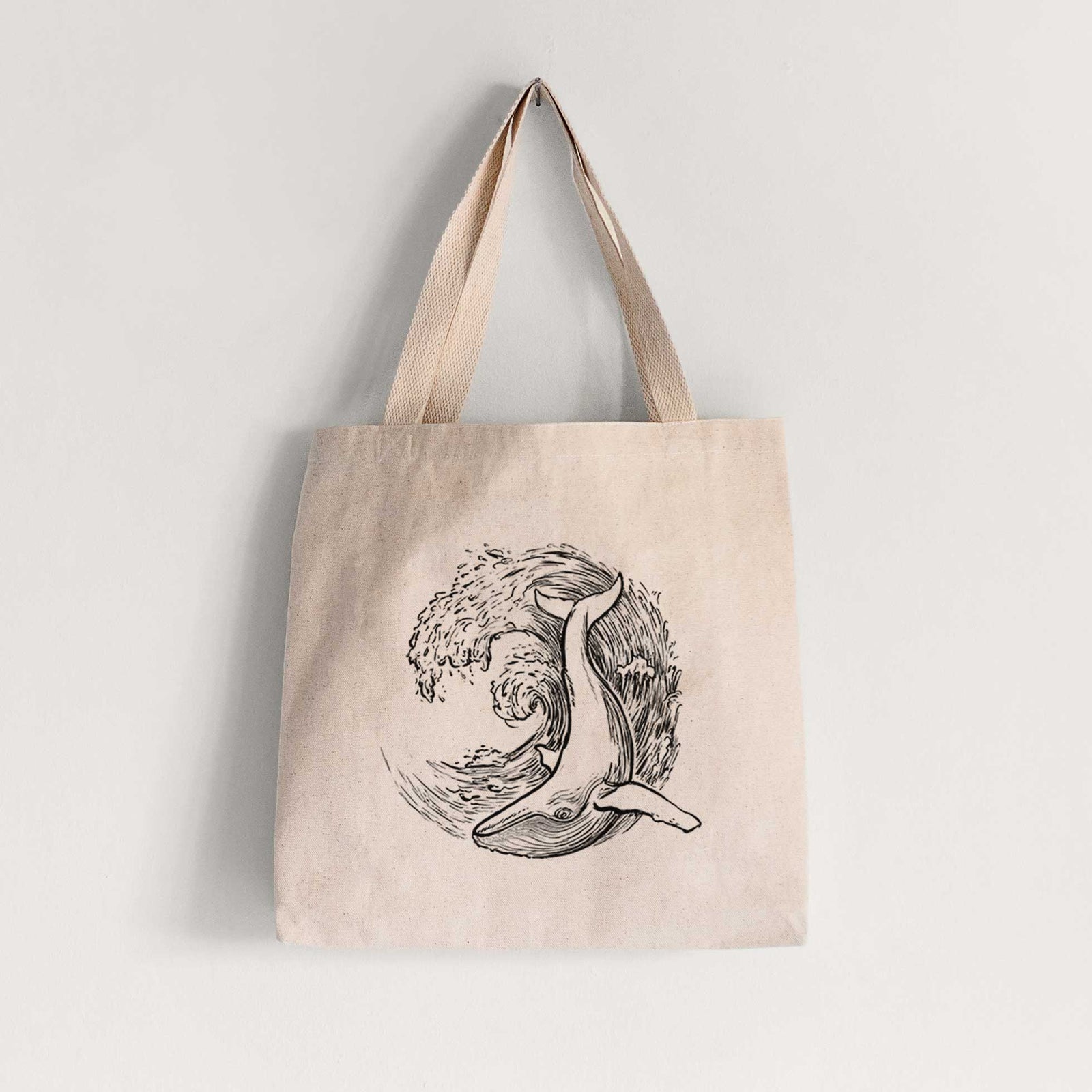 Natural canvas tote bag printed with a black ink illustration of a humpback whale swimming inside a curling ocean wave.