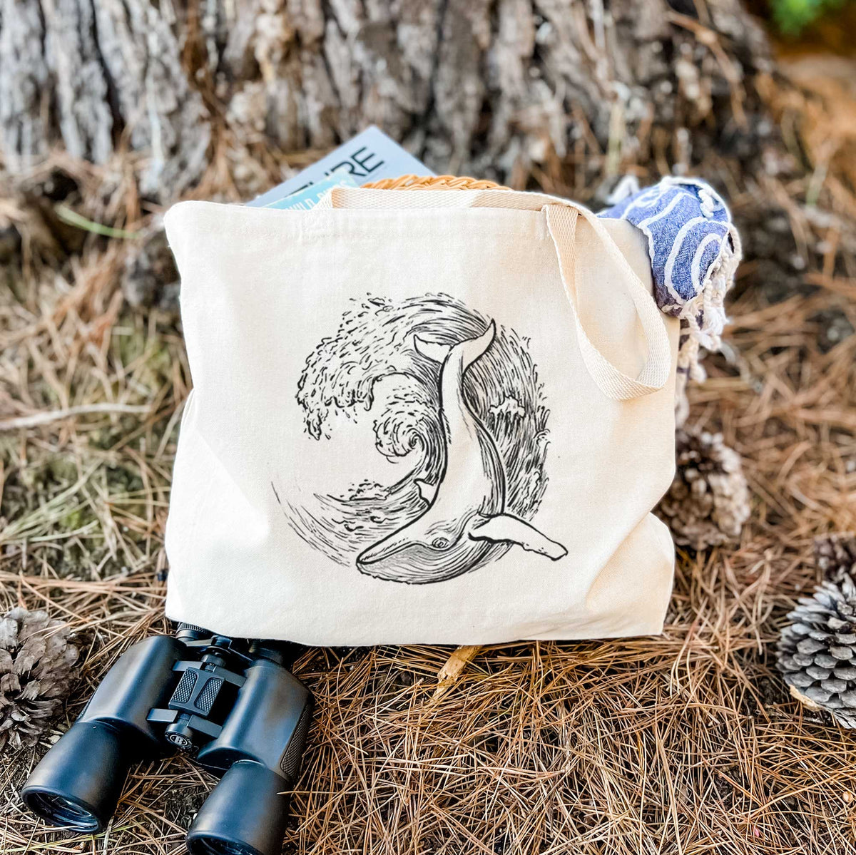 Canvas tote bag printed with a black ink illustration of a humpback whale swimming inside a curling ocean wave, resting on dry grass beside a pair of binoculars.
