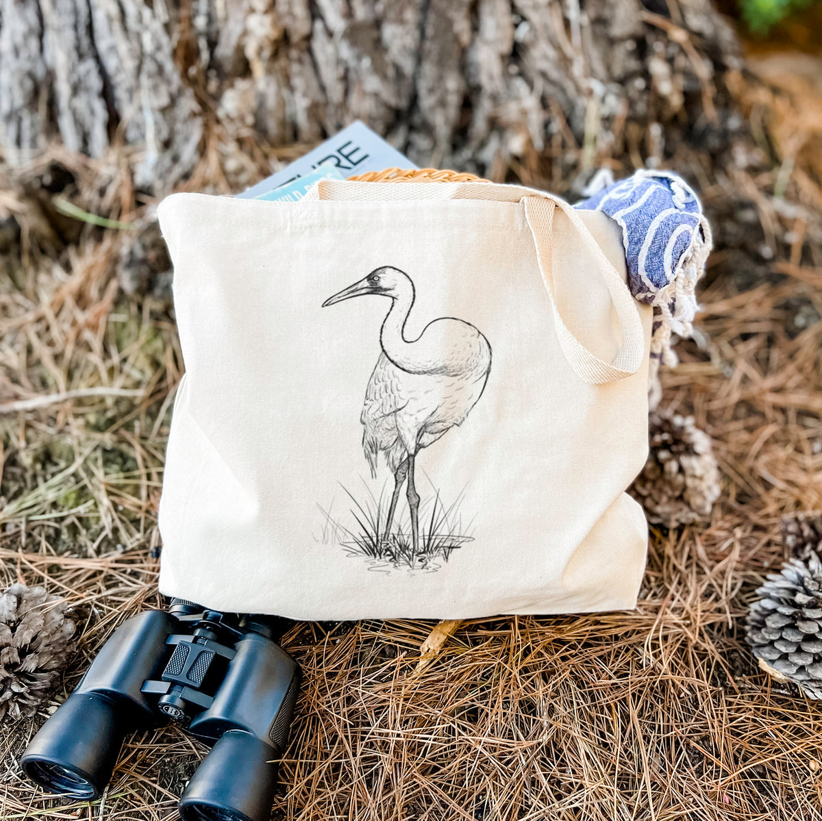 Canvas tote bag printed with a detailed black ink illustration of a whooping crane standing in grass, resting on dry grass beside a pair of binoculars.
