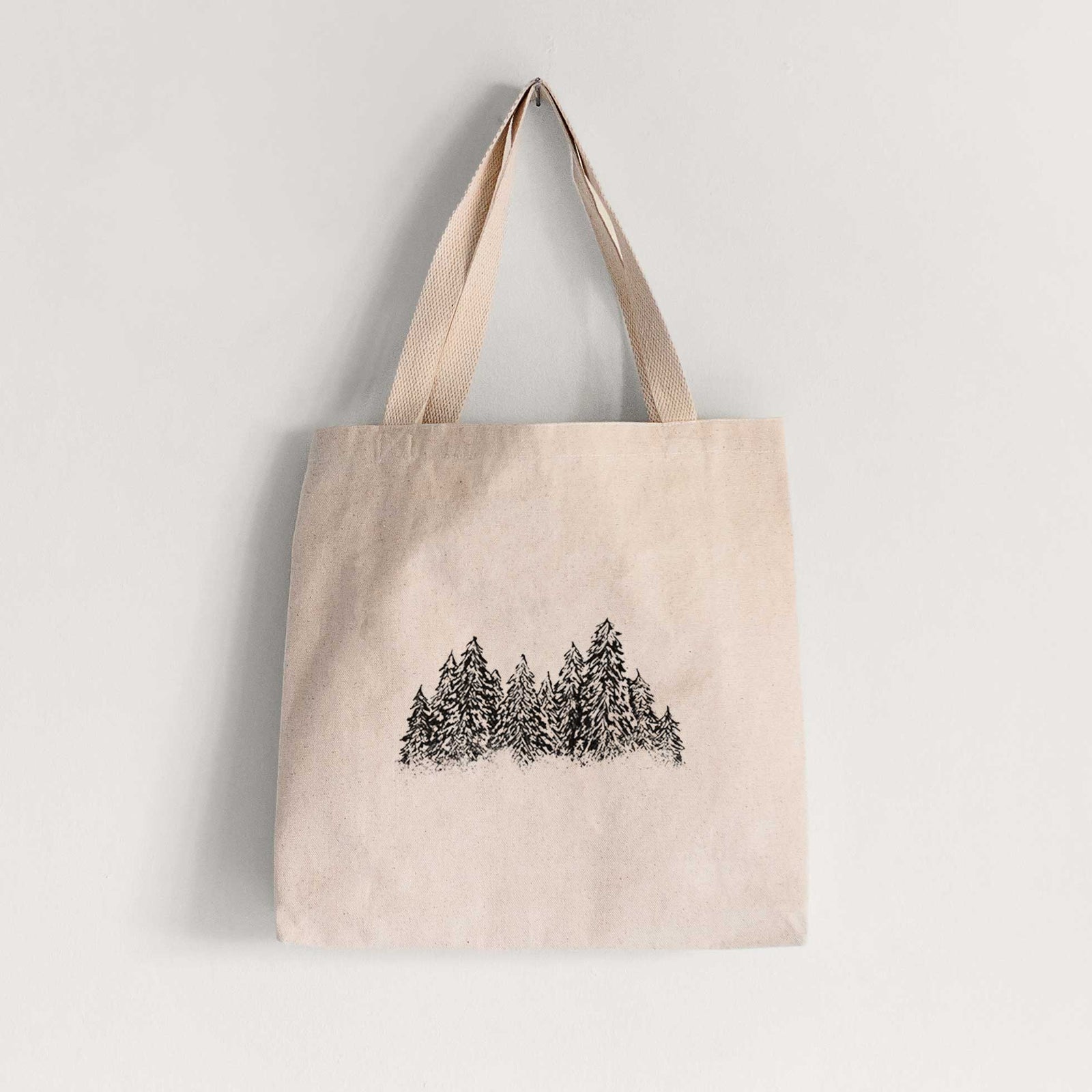 Natural canvas tote bag printed with a black ink illustration of a row of snow-dusted evergreen trees.