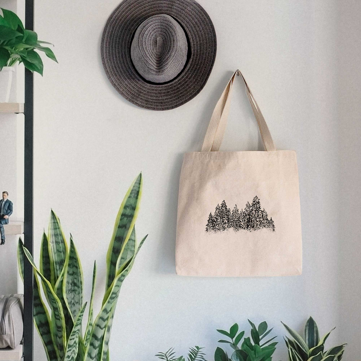 Canvas tote bag printed with a black ink illustration of a row of snow-dusted evergreen trees, hanging on a wall hook below a hat, next to potted plants.