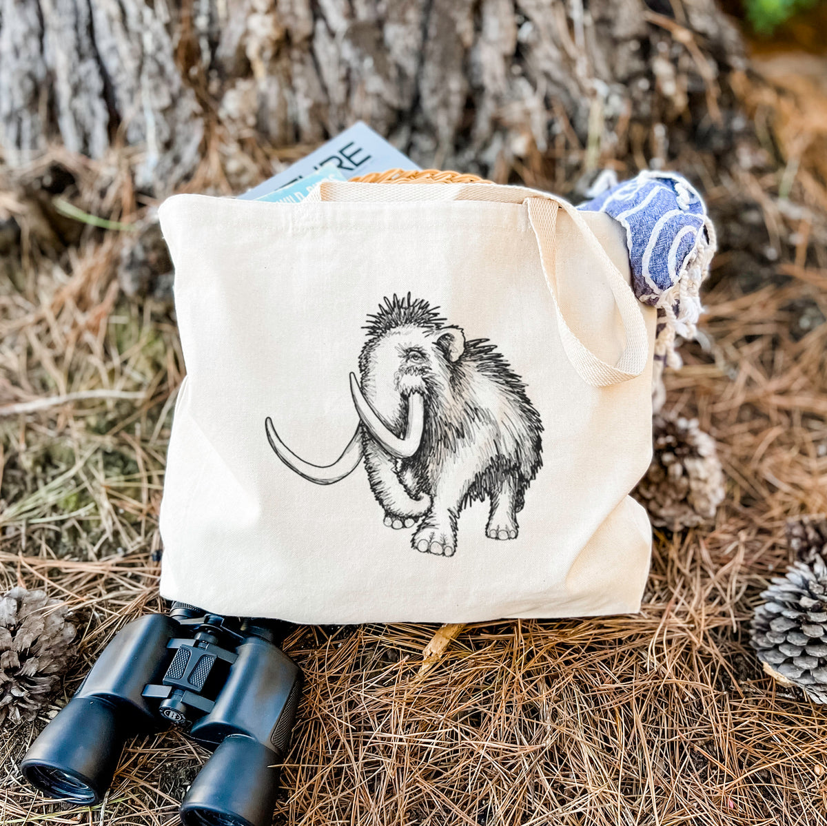 Canvas tote bag printed with a black ink illustration of a woolly mammoth with long curved tusks, resting on dry grass beside a pair of binoculars.