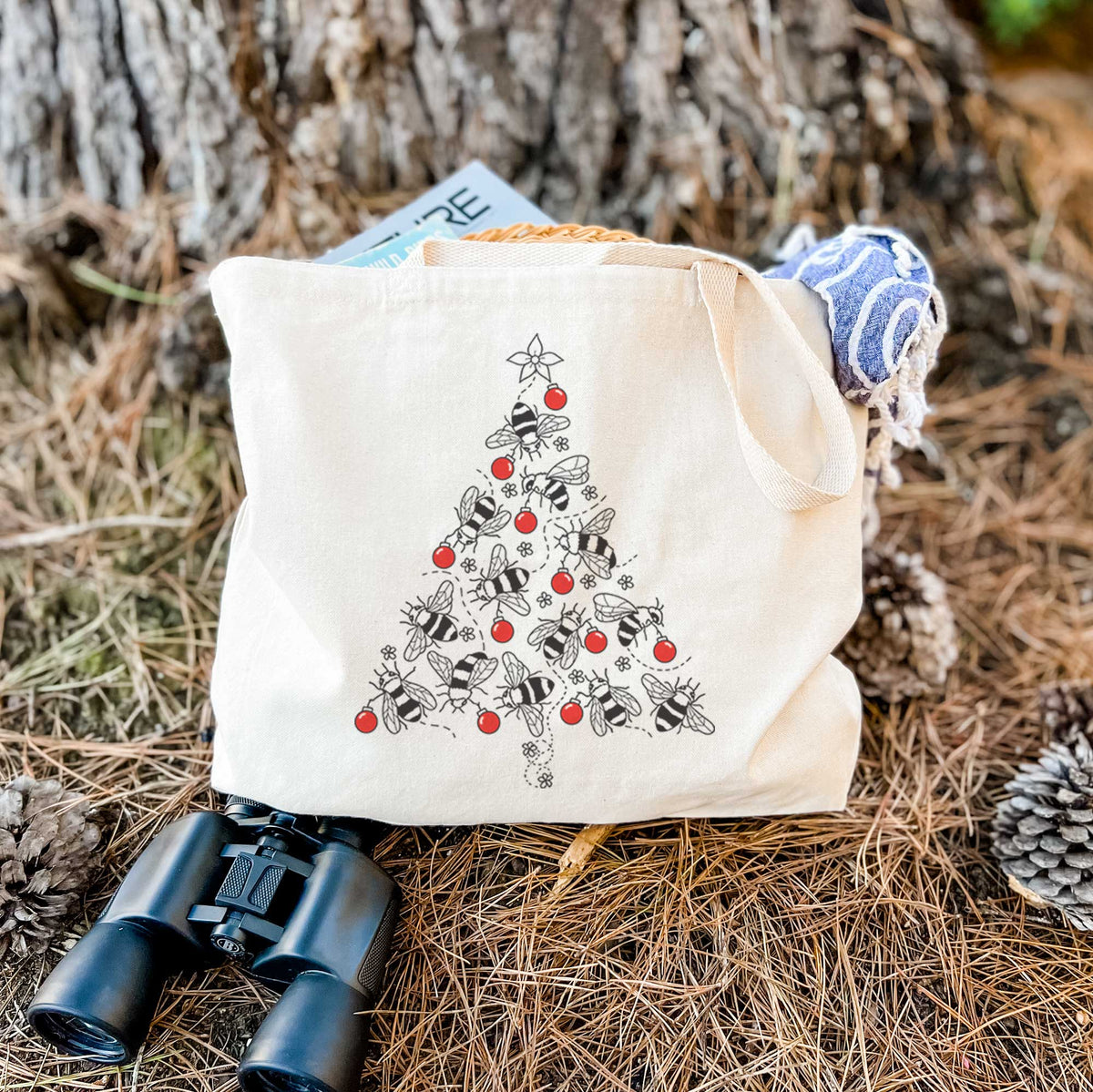 Canvas tote bag printed with a Christmas tree made of black ink bees with red ornaments and a star on top, resting on dry grass beside a pair of binoculars.