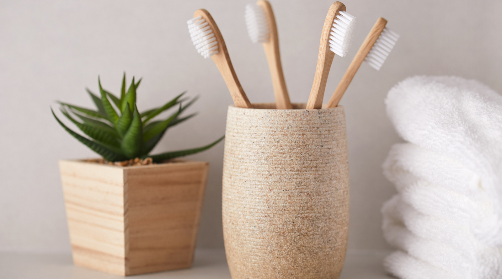 Bamboo toothbrushes in a ceramic holder next to a small potted plant and white towels, illustrating how to use less plastic in personal care items