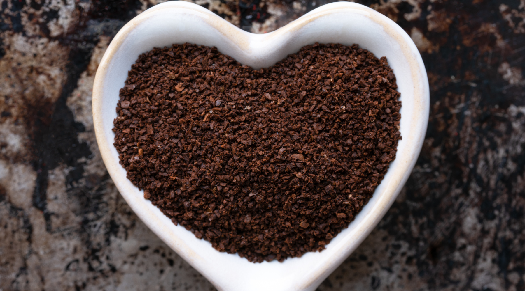 Heart-shaped bowl filled with coffee grounds on a rustic surface, illustrating how to use coffee grounds in garden for soil enrichment and plant growth.