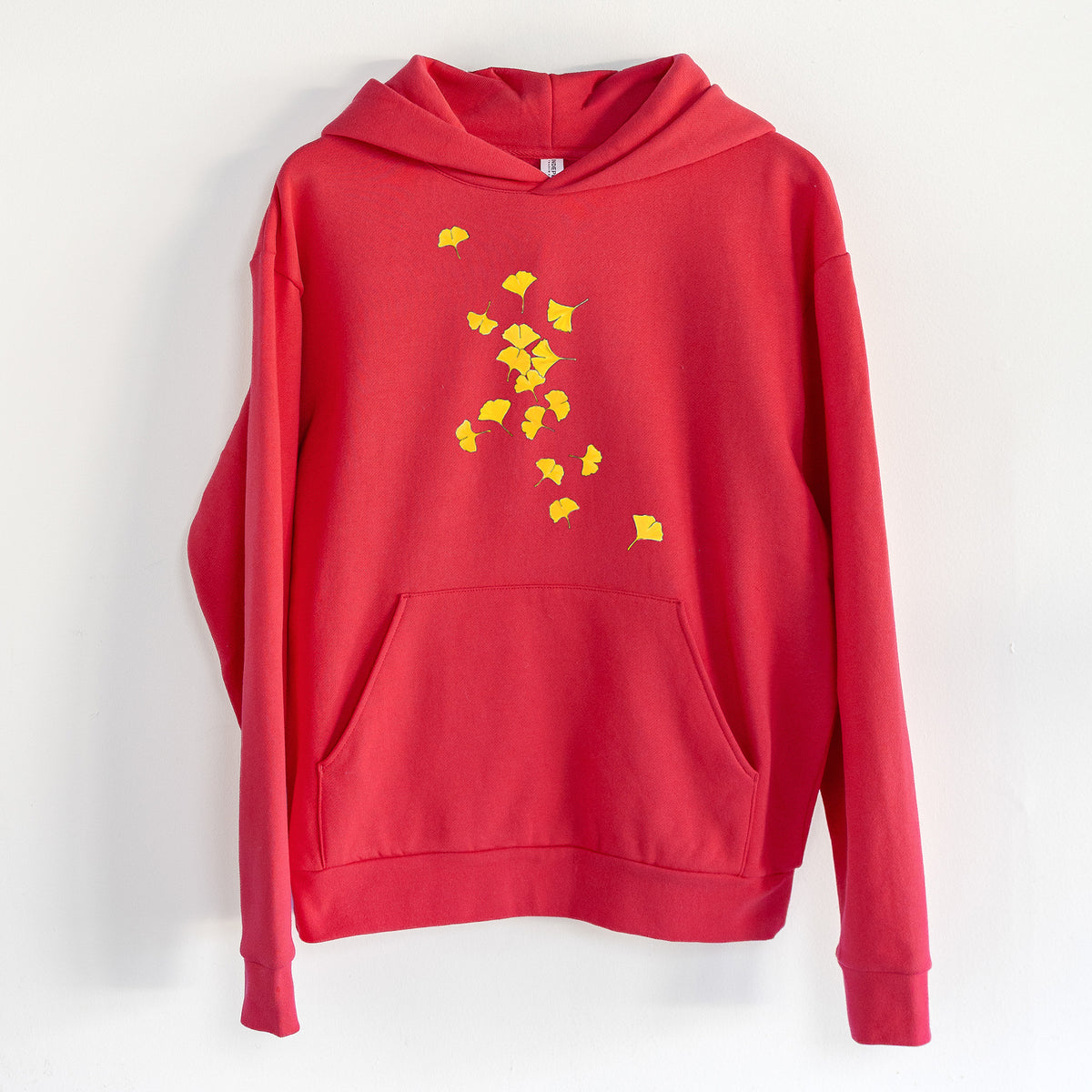 Falling Ginkgo Leaves - Statement Midweight Hoodie in Cherry Red, hanging on a white wall, with a front kangaroo pocket and no drawcords.